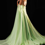 A woman modeling a flowing light green evening gown with thin straps and a long train, photographed against a black studio background. The brand name RACHEL ALLAN appears in the top right corner.