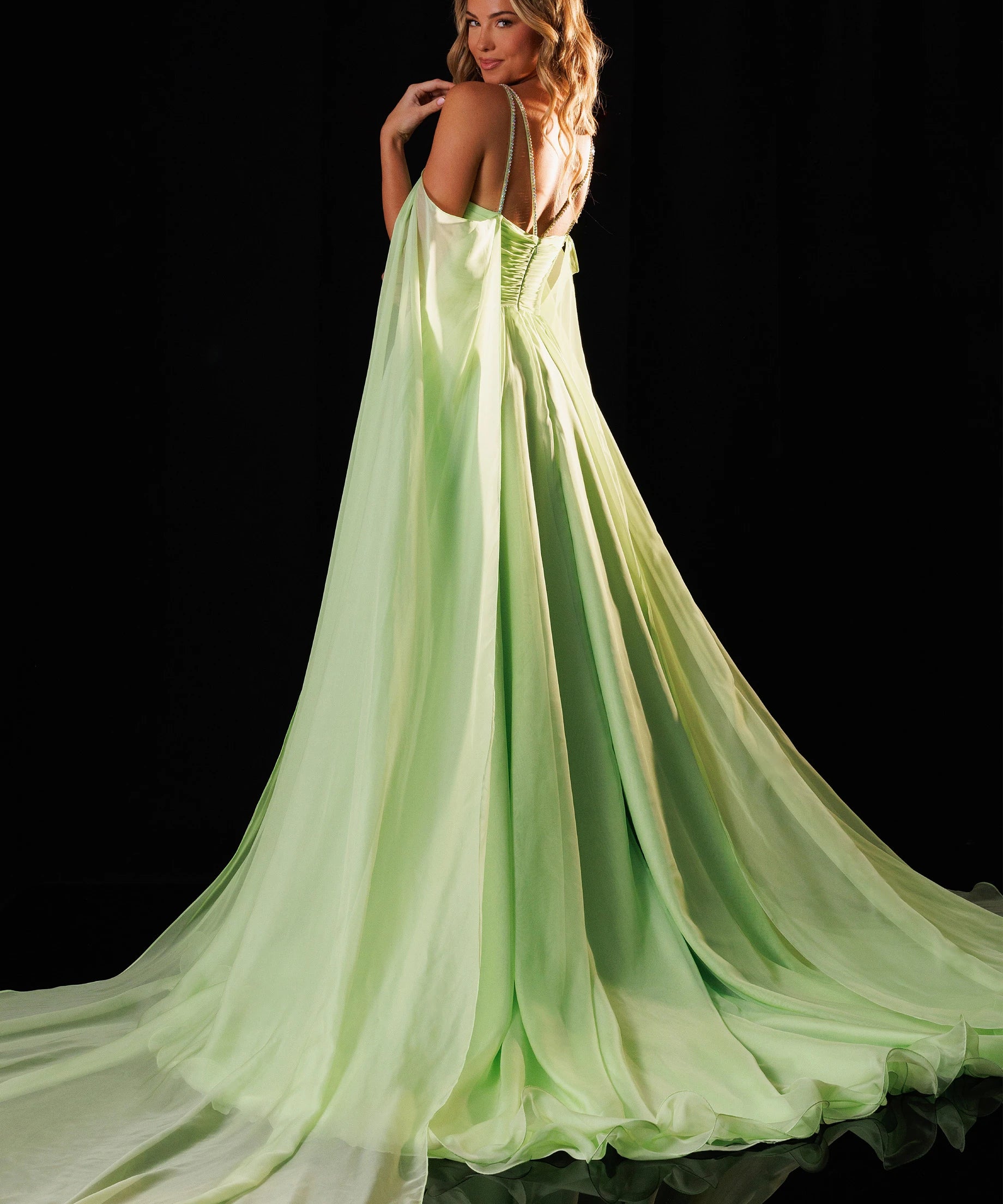 A woman modeling a flowing light green evening gown with thin straps and a long train, photographed against a black studio background. The brand name RACHEL ALLAN appears in the top right corner.
