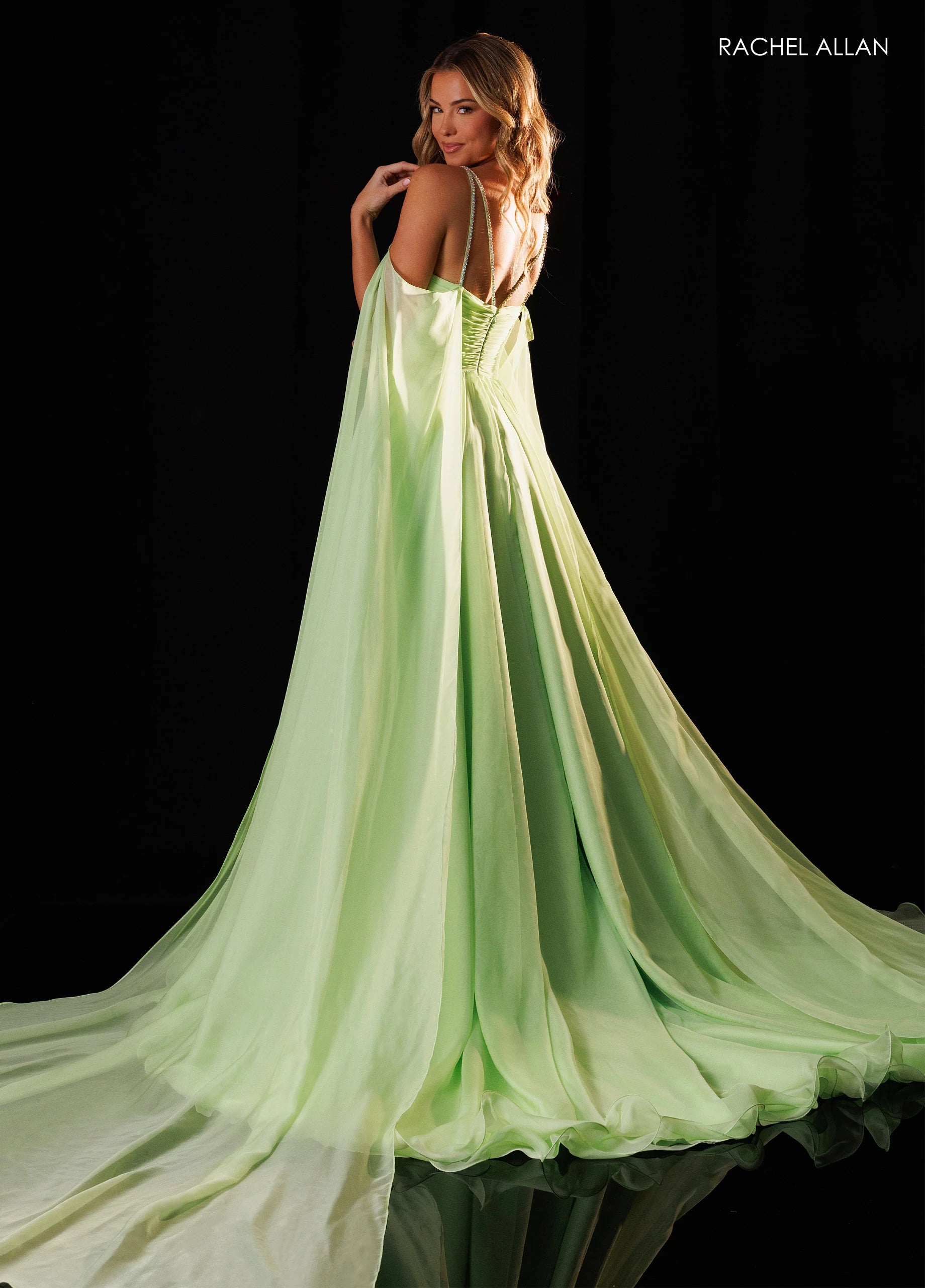 A woman modeling a flowing light green evening gown with thin straps and a long train, photographed against a black studio background. The brand name RACHEL ALLAN appears in the top right corner.