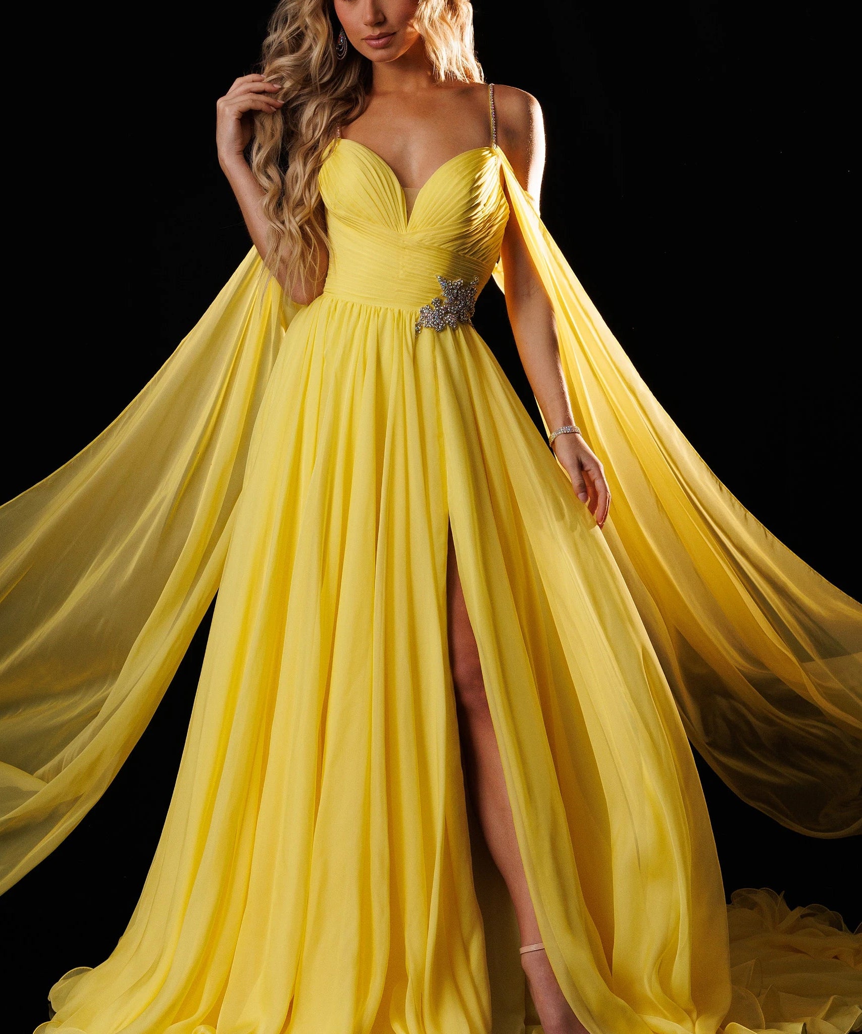 A woman models a bright yellow formal gown with a sweetheart neckline, thin straps, a high thigh slit, and a flowing sheer cape train against a black background. The brand name 'RACHEL ALLAN' is printed in the top right corner.