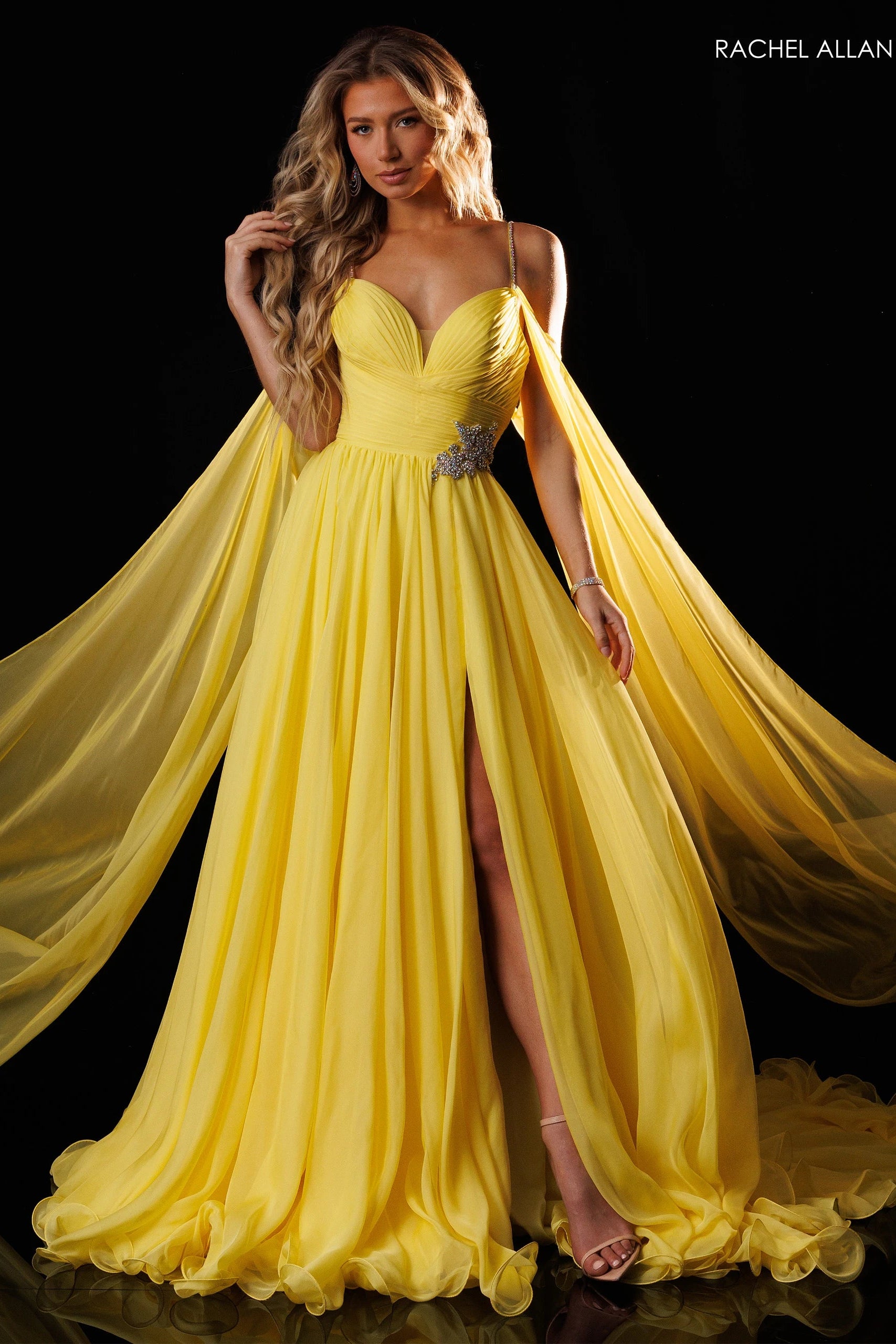 A woman models a bright yellow formal gown with a sweetheart neckline, thin straps, a high thigh slit, and a flowing sheer cape train against a black background. The brand name 'RACHEL ALLAN' is printed in the top right corner.