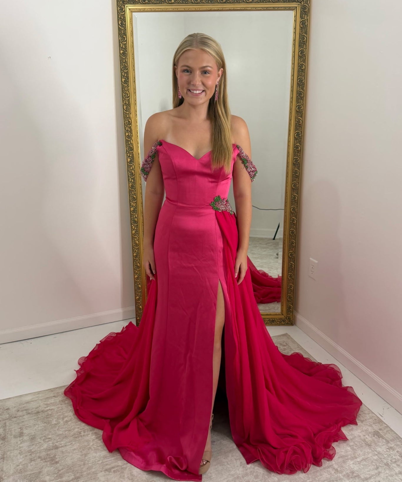 A woman stands in front of a large gold-framed mirror wearing a bright fuchsia off-the-shoulder evening gown with a thigh-high slit and a dramatic train in a room with white walls and a light rug.