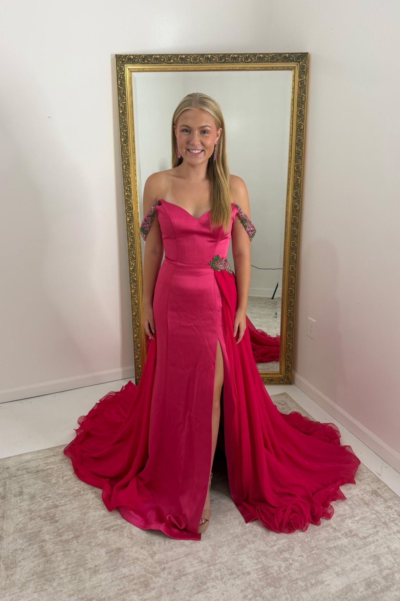 A woman stands in front of a large gold-framed mirror wearing a bright fuchsia off-the-shoulder evening gown with a thigh-high slit and a dramatic train in a room with white walls and a light rug.