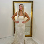 A woman stands indoors wearing a fitted lace gown with a sheer draped train, posed in front of a large gold-framed floor mirror against white walls.