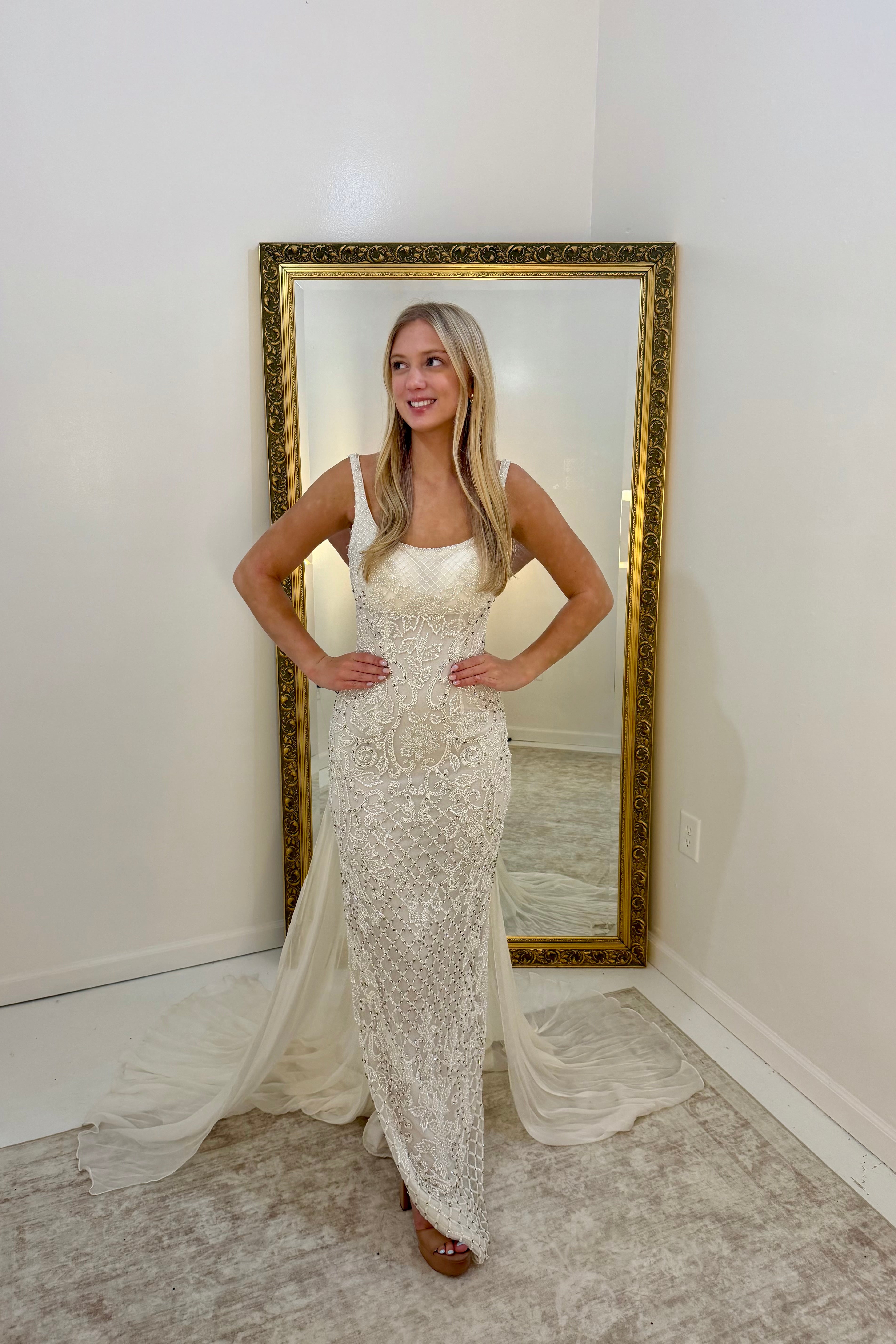 A woman stands indoors wearing a fitted lace gown with a sheer draped train, posed in front of a large gold-framed floor mirror against white walls.