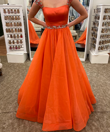 An orange formal ball gown with a square neckline, embellished cap sleeves, and a beaded waistband, shown on a model in a clothing boutique.