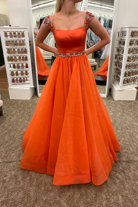 An orange formal ball gown with a square neckline, embellished cap sleeves, and a beaded waistband, shown on a model in a clothing boutique.