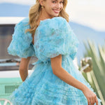 A woman wearing a light blue ruffled tulle dress with oversized puff sleeves, photographed outdoors in front of a vintage-style vehicle and plants. The brand name 'SHERRI HILL' appears in the bottom right corner.