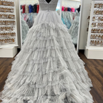 A voluminous silver-gray tulle ball gown displayed on a black mannequin torso inside a bridal boutique. The dress features a deep V-neckline, a fitted bodice, and a heavily layered, ruffled tulle skirt that cascades to the floor.