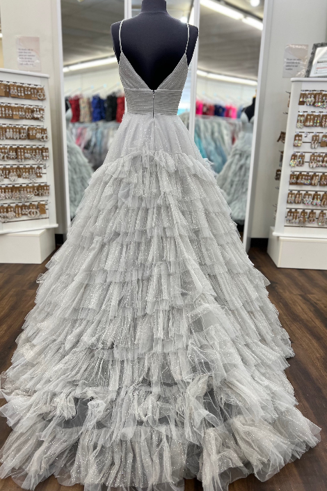 A voluminous silver-gray tulle ball gown displayed on a black mannequin torso inside a bridal boutique. The dress features a deep V-neckline, a fitted bodice, and a heavily layered, ruffled tulle skirt that cascades to the floor.