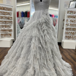 A voluminous white ball gown with a sweetheart neckline and a heavily ruffled, tiered tulle skirt, displayed on a black mannequin in a bridal boutique.
