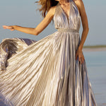 A woman wearing a metallic champagne-silver halter maxi dress with a pleated skirt, standing in shallow water on a beach with the dress flowing in the wind. The Sherri Hill brand name is visible in the bottom right corner.