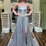 A woman modeling a strapless silver formal gown with a pleated skirt, high leg slit, and a shimmering embellished waistband, standing in a boutique.