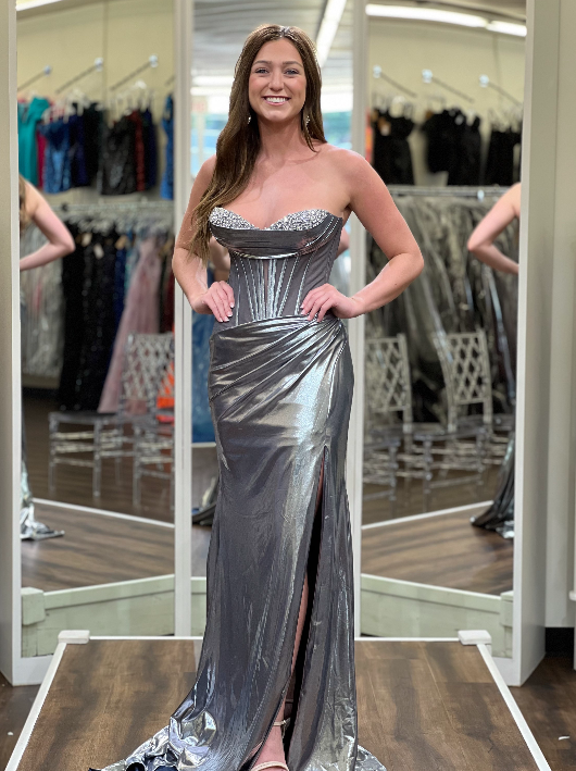 A woman modeling a strapless metallic silver evening gown with a sweetheart neckline, beaded bodice, and thigh-high slit in a fitting room with mirrors and dress racks.