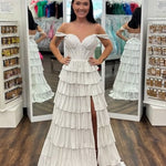 A woman modeling a white off-the-shoulder tiered gown with a thigh-high slit, standing in front of a full-length mirror inside a boutique.