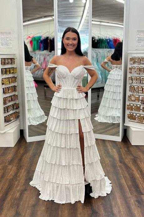 A woman modeling a white off-the-shoulder tiered gown with a thigh-high slit, standing in front of a full-length mirror inside a boutique.
