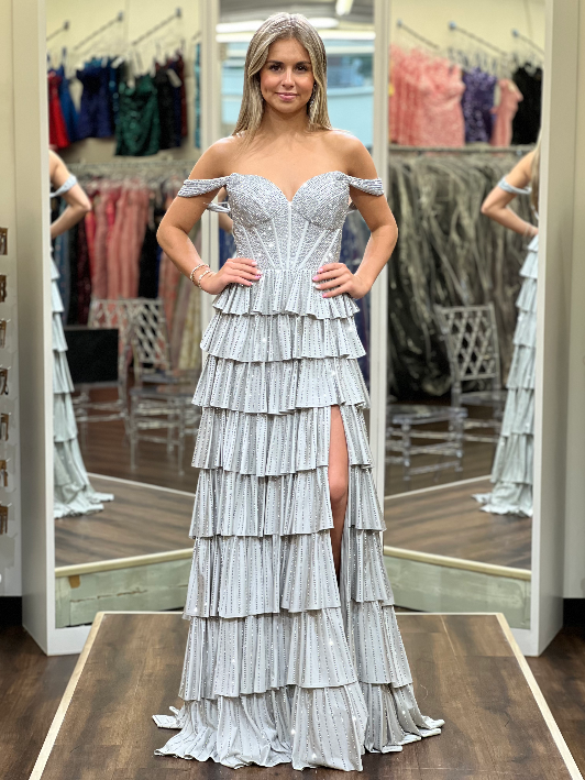 A woman models a floor-length off-the-shoulder gown with a fitted embellished bodice and dramatic tiered ruffle skirt in a bridal or formal salon setting with mirrors and racks of dresses in the background.