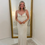 A woman wearing a thin strap, floor-length champagne-colored gown with vertical bead and sequin stripes, standing in front of a large gold-framed floor mirror in a white room.