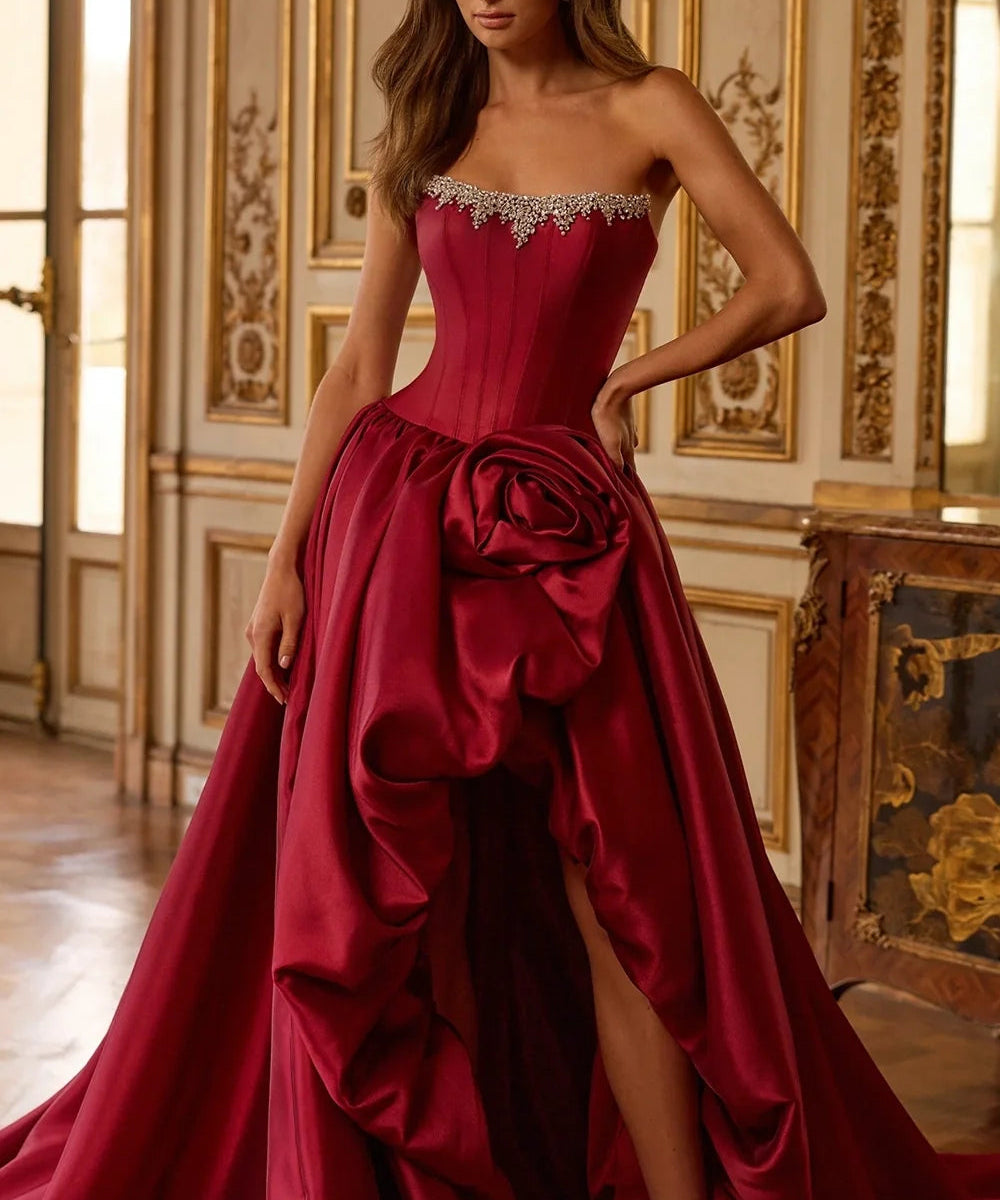 A woman models a strapless formal gown in a luxurious interior room with gold-trimmed walls. The gown is a rich deep red with a structured corset bodice, embellished sweetheart neckline, a large sculptural rose detail at the waist, and a high-slit skirt with a long train.