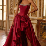 A woman models a strapless formal gown in a luxurious interior room with gold-trimmed walls. The gown is a rich deep red with a structured corset bodice, embellished sweetheart neckline, a large sculptural rose detail at the waist, and a high-slit skirt with a long train.