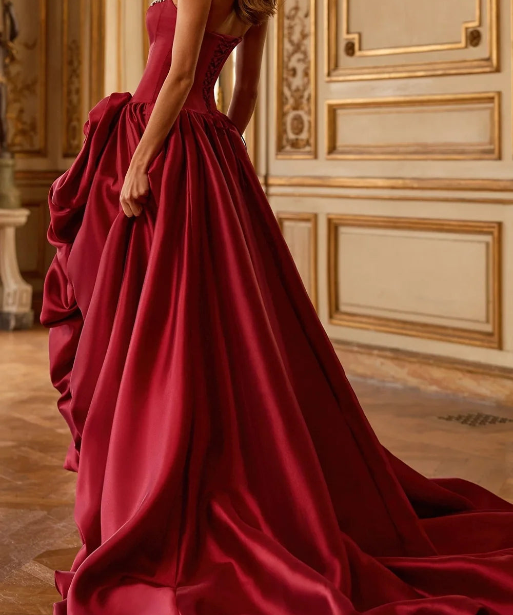 A woman wearing a floor-length burgundy satin ball gown with a structured corset bodice and dramatic ruffled overskirt, standing in an ornate cream and gold interior room.