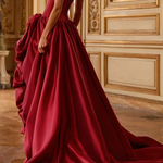 A woman wearing a floor-length burgundy satin ball gown with a structured corset bodice and dramatic ruffled overskirt, standing in an ornate cream and gold interior room.
