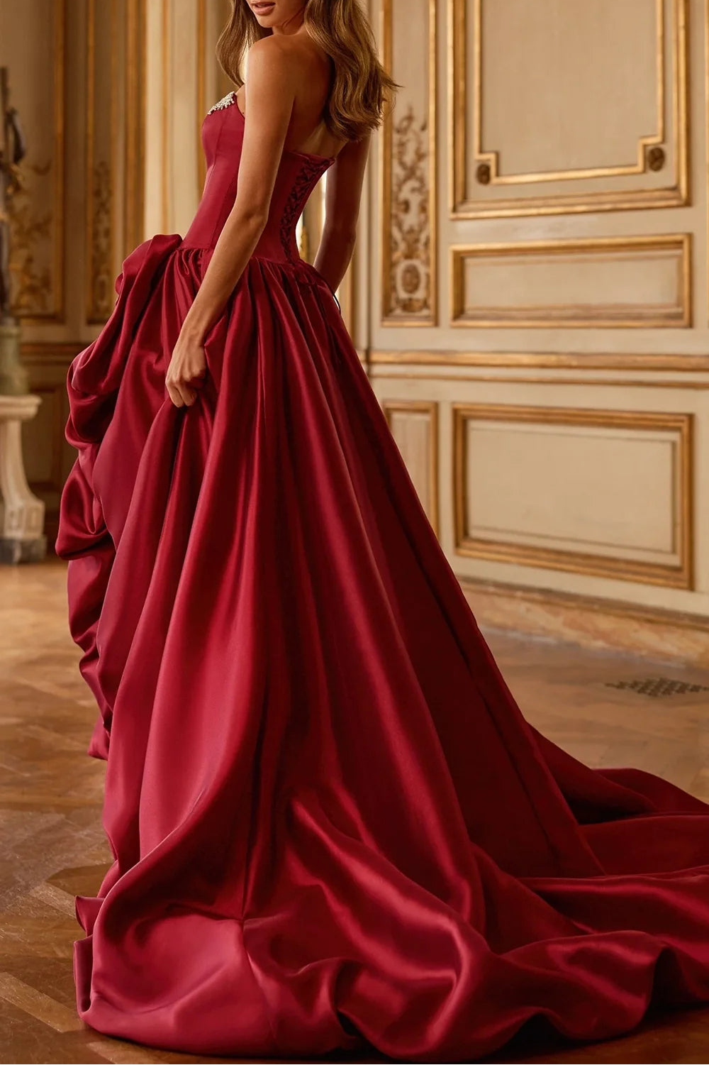 A woman wearing a floor-length burgundy satin ball gown with a structured corset bodice and dramatic ruffled overskirt, standing in an ornate cream and gold interior room.