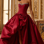 A woman models a strapless ball gown in deep burgundy satin, featuring a structured corset bodice, a large sculptural rose detail at the waist, and a dramatic overskirt, set against an ornate, warm-toned interior room.