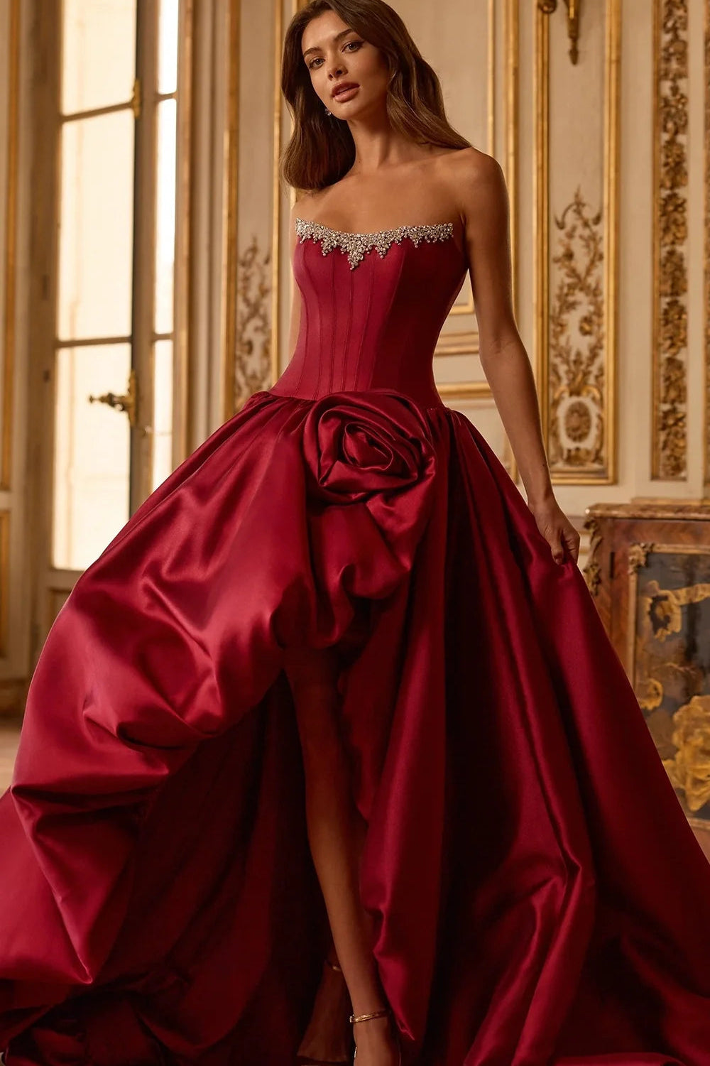 A woman models a strapless ball gown in deep burgundy satin, featuring a structured corset bodice, a large sculptural rose detail at the waist, and a dramatic overskirt, set against an ornate, warm-toned interior room.