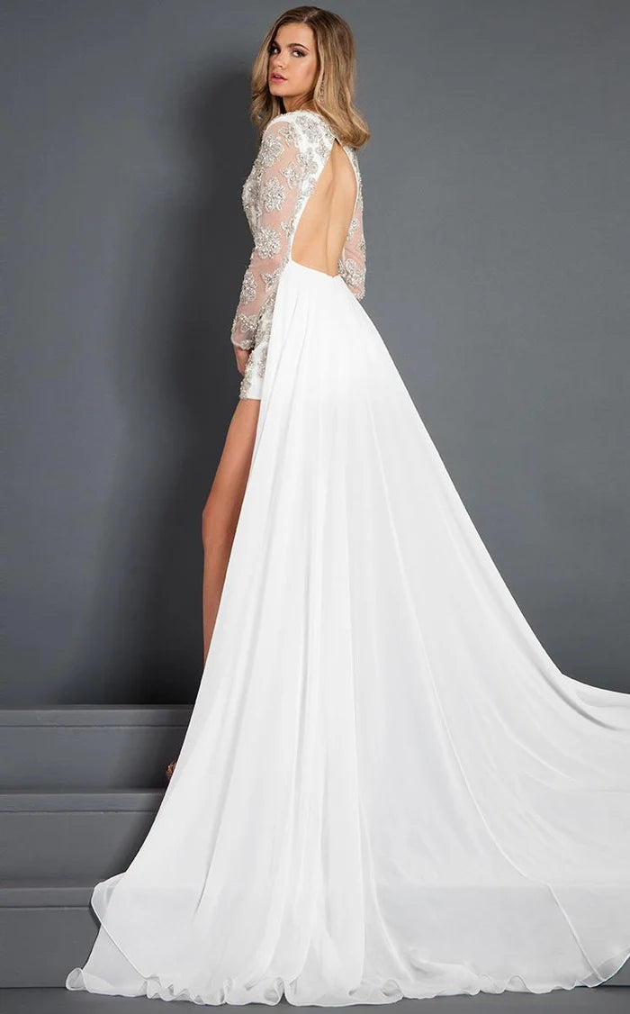 A woman modeling a white bridal gown with a sheer lace long-sleeve backless bodice, high slit, and a flowing train against a dark gray studio background.