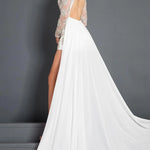 A woman modeling a white bridal gown with a sheer lace long-sleeve backless bodice, high slit, and a flowing train against a dark gray studio background.