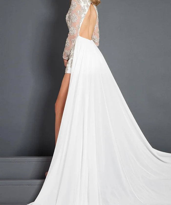 A woman modeling a white bridal gown with a sheer lace long-sleeve backless bodice, high slit, and a flowing train against a dark gray studio background.