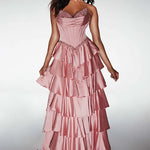A woman modeling a strapless formal gown with a structured corset-style bodice and a full, tiered satin skirt in a dusty pink color, set against a light gray studio background.