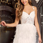 A woman modeling a short white dress with a lace bustier-style bodice and a fluffy feathered skirt, posed in front of a large industrial vault door. The Sherri Hill brand name is visible in the bottom right corner.