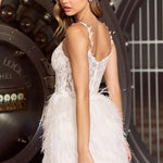 A woman modeling a short white lace and feather dress, photographed from the back in front of a large industrial vault door. The dress features a lace bodice with spaghetti straps and a voluminous white feathered skirt.