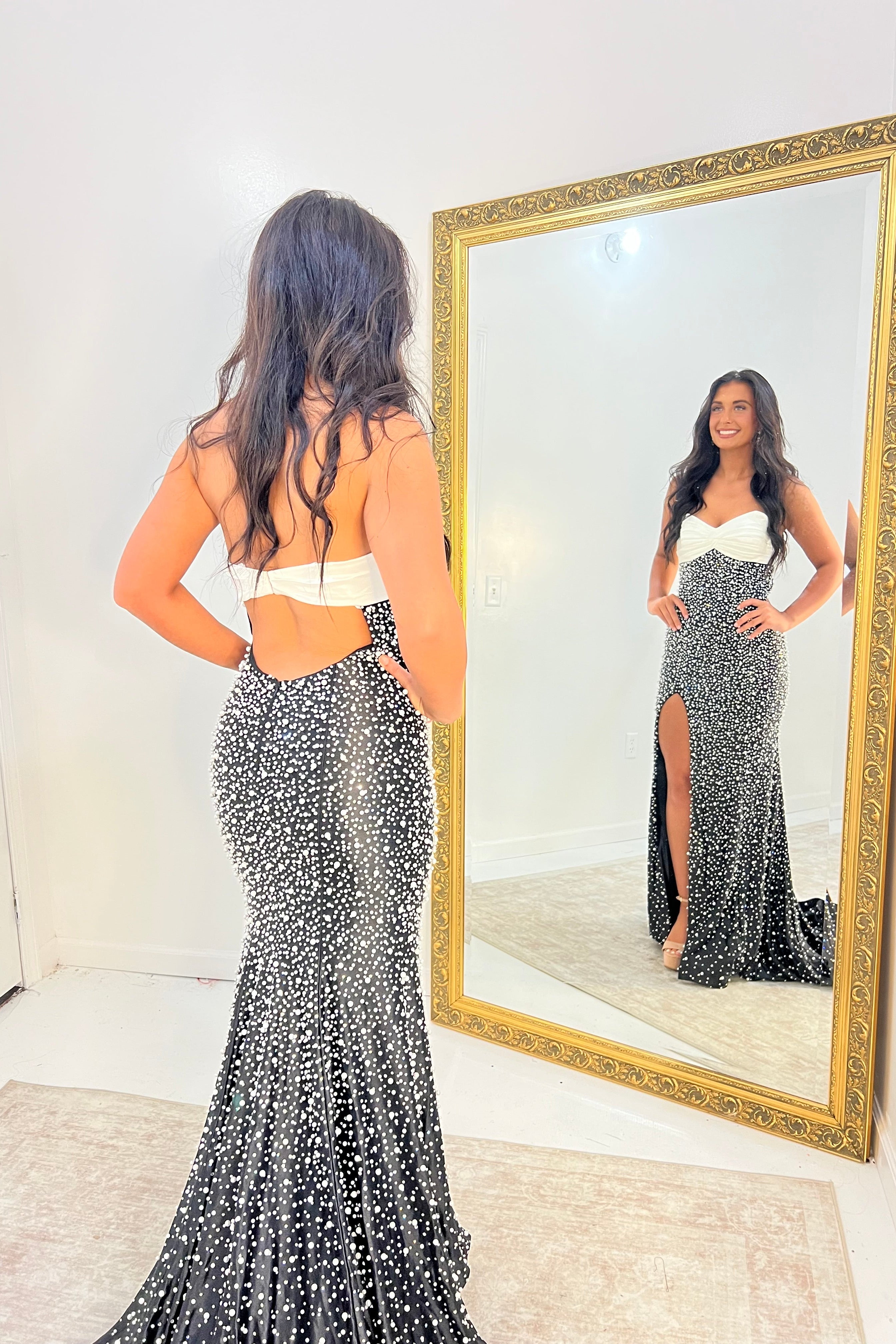 A woman stands in a room wearing a sparkling black and silver mermaid gown with a white sweetheart top, facing a large gold-framed mirror that shows her front view. The dress has a high slit and an open back.