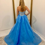 A woman stands with her back to the camera wearing a strapless light blue ball gown with a beaded corset-style bodice and a voluminous tulle skirt, posing in front of a large gold-framed mirror.