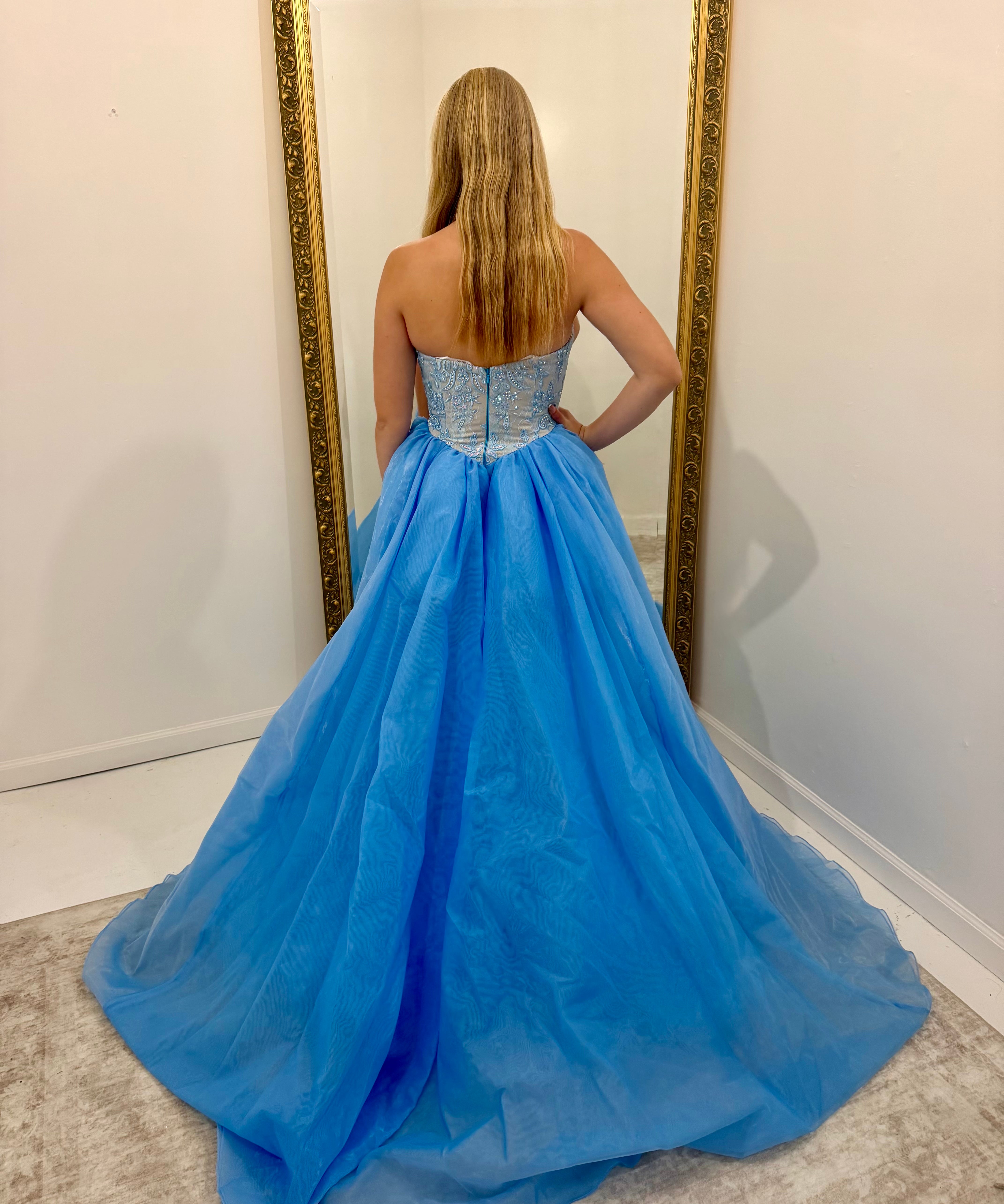 A woman stands with her back to the camera wearing a strapless light blue ball gown with a beaded corset-style bodice and a voluminous tulle skirt, posing in front of a large gold-framed mirror.