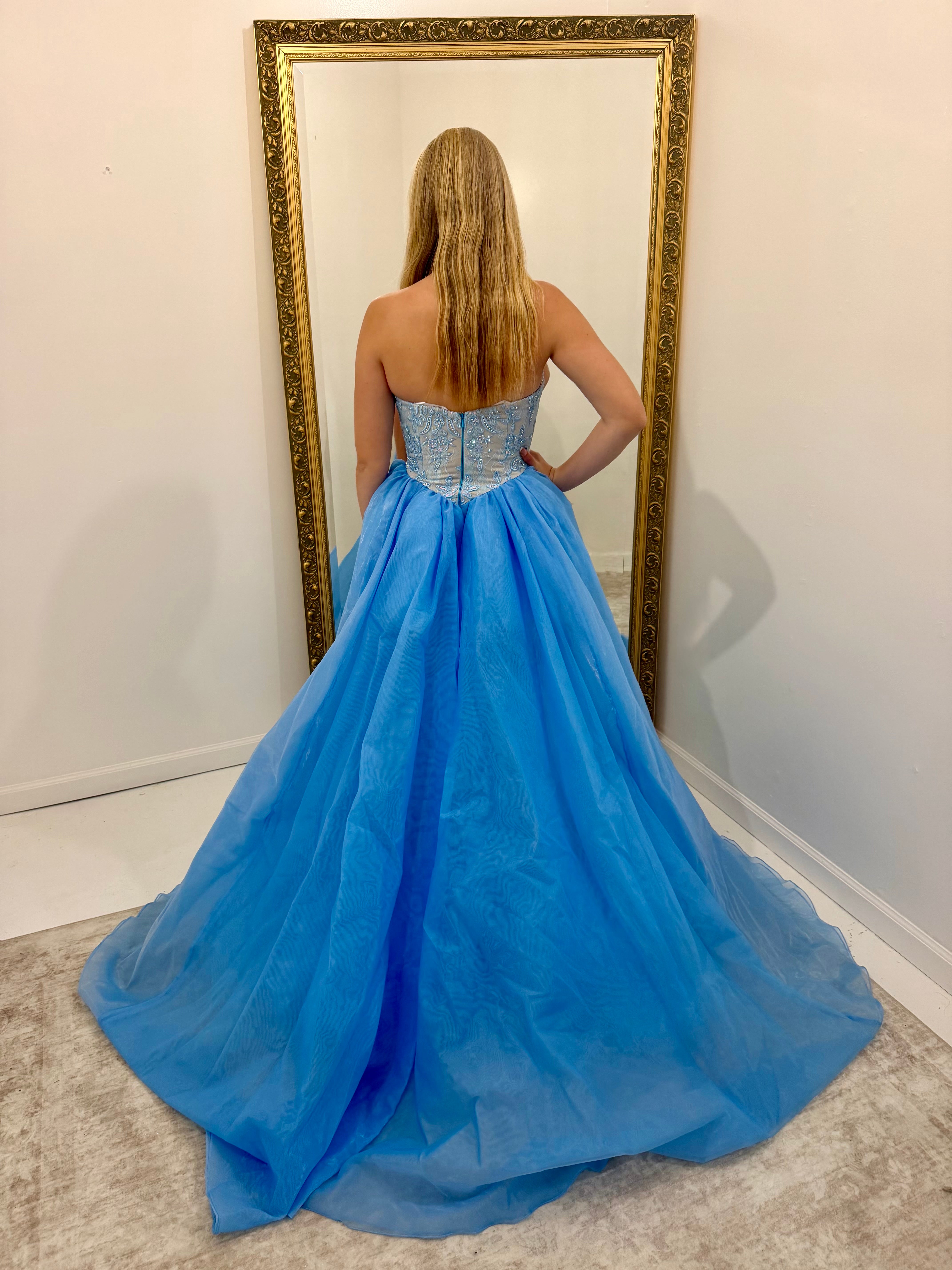 A woman stands with her back to the camera wearing a strapless light blue ball gown with a beaded corset-style bodice and a voluminous tulle skirt, posing in front of a large gold-framed mirror.