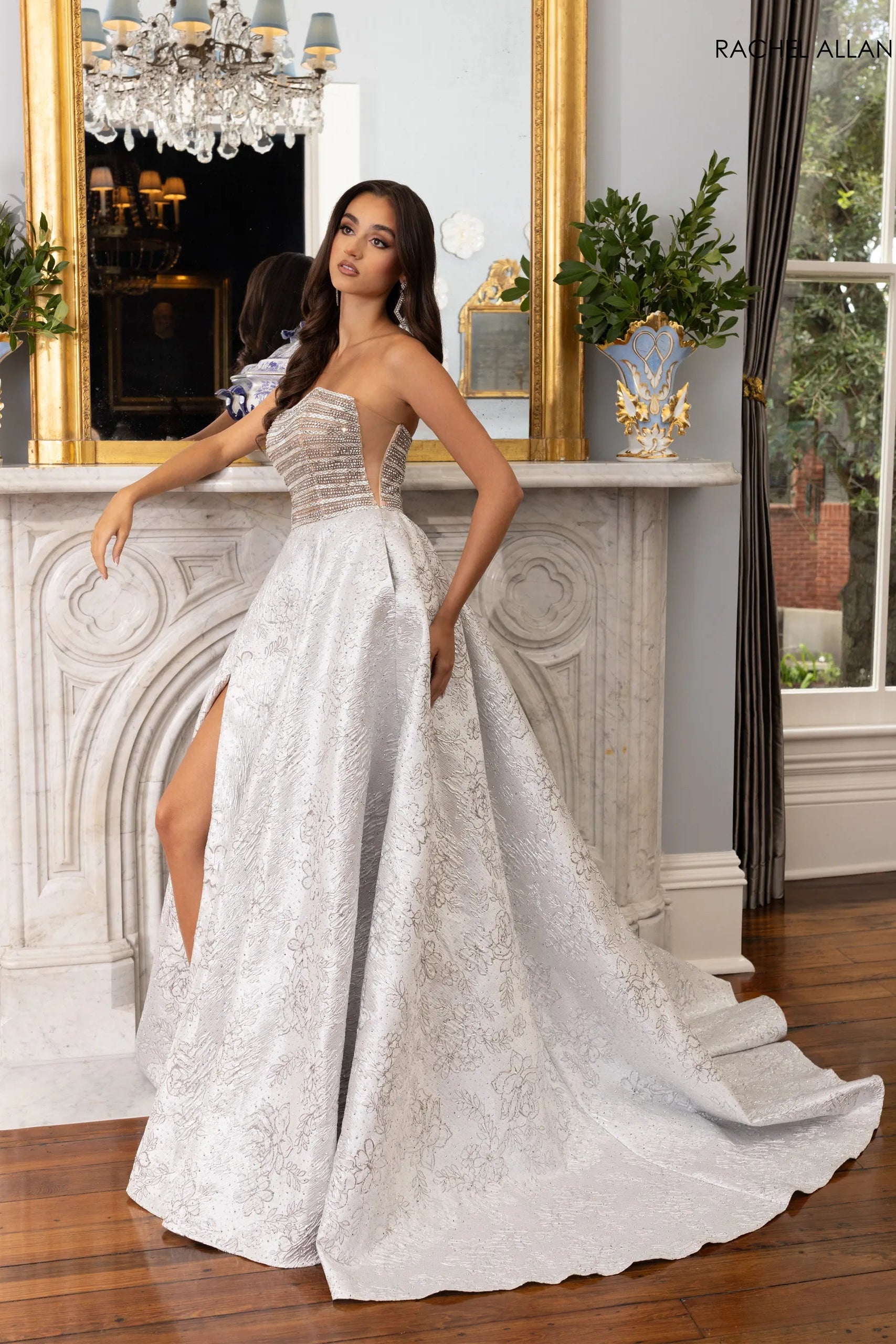A woman models a strapless white lace ball gown with a sweetheart neckline, sheer corset-style bodice, high leg slit, and a long train in an elegant indoor room with a fireplace and mirror.