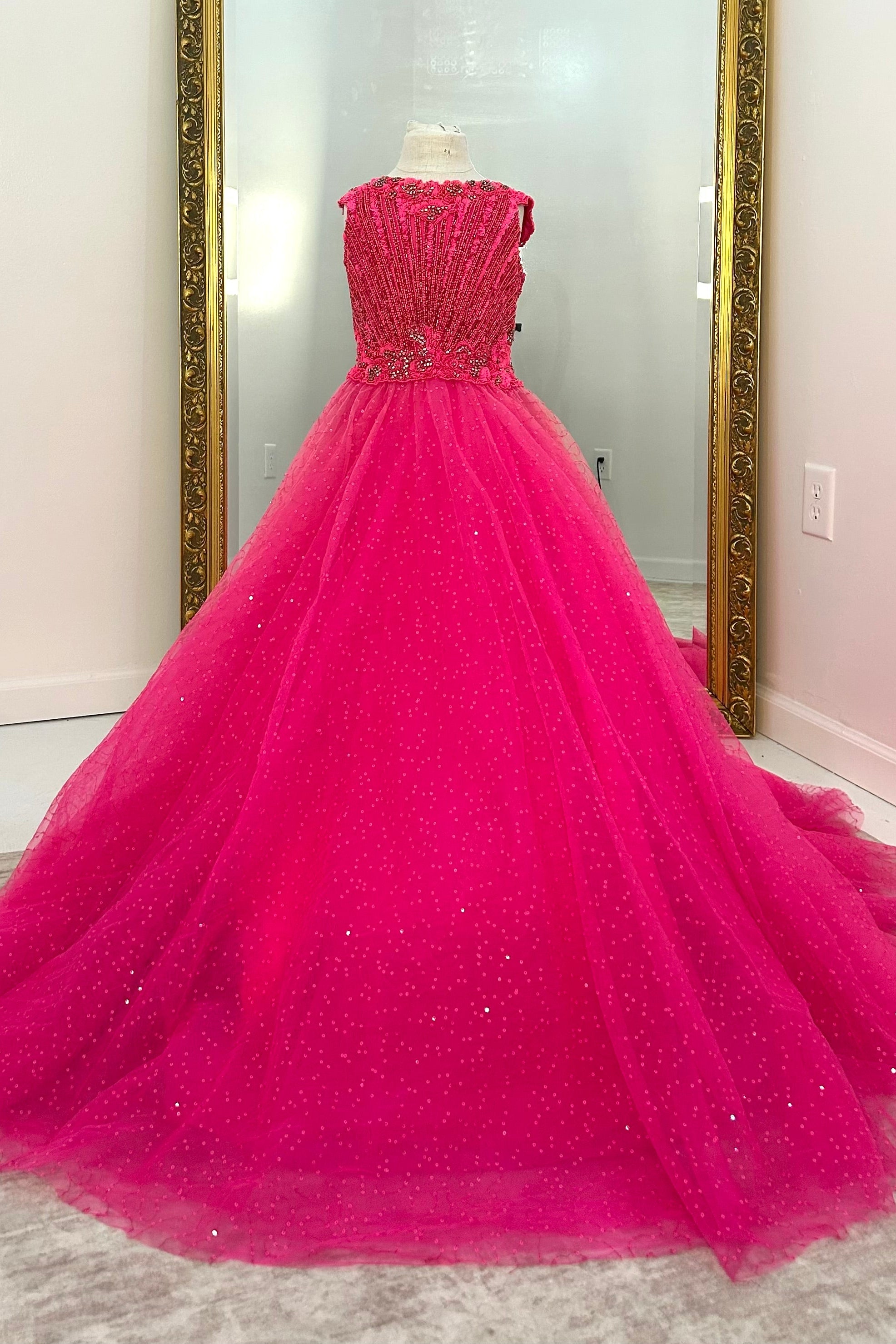 A bright fuchsia pink ball gown with a beaded strapless bodice and a voluminous tulle skirt, displayed on a dress form in an indoor room with mirrors.