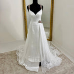 A white sleeveless gown displayed on a black mannequin in front of a large gold-framed mirror, with a long train spread across a light beige carpeted floor.