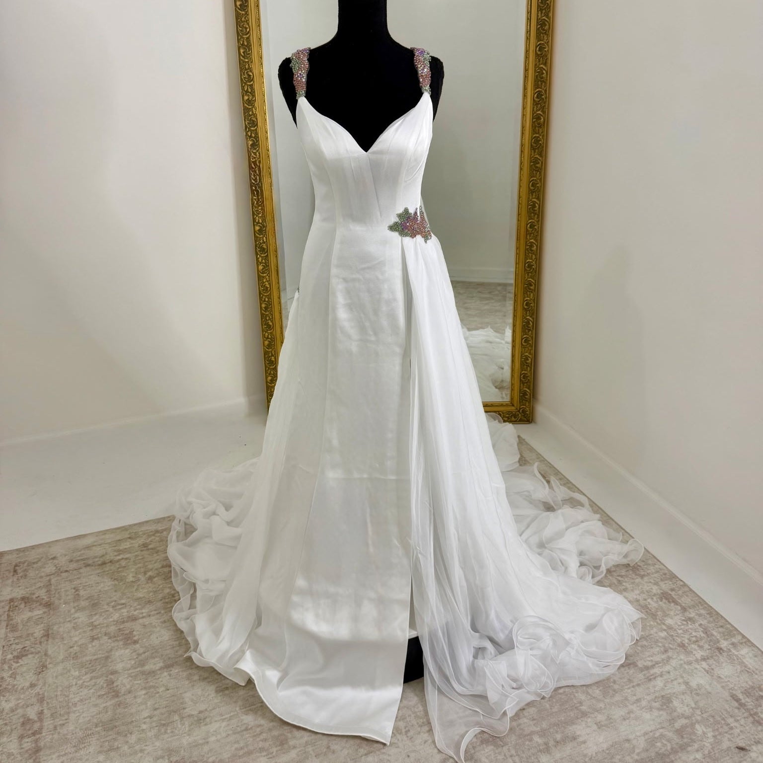 A white sleeveless gown displayed on a black mannequin in front of a large gold-framed mirror, with a long train spread across a light beige carpeted floor.