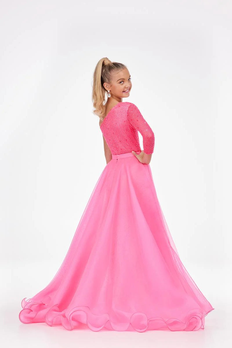 A young girl with blonde hair in a ponytail is shown from the back wearing a bright pink formal gown with a lace bodice and a flowing tulle skirt, set against a plain white studio background.