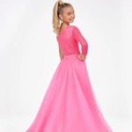 A young girl with blonde hair in a ponytail is shown from the back wearing a bright pink formal gown with a lace bodice and a flowing tulle skirt, set against a plain white studio background.