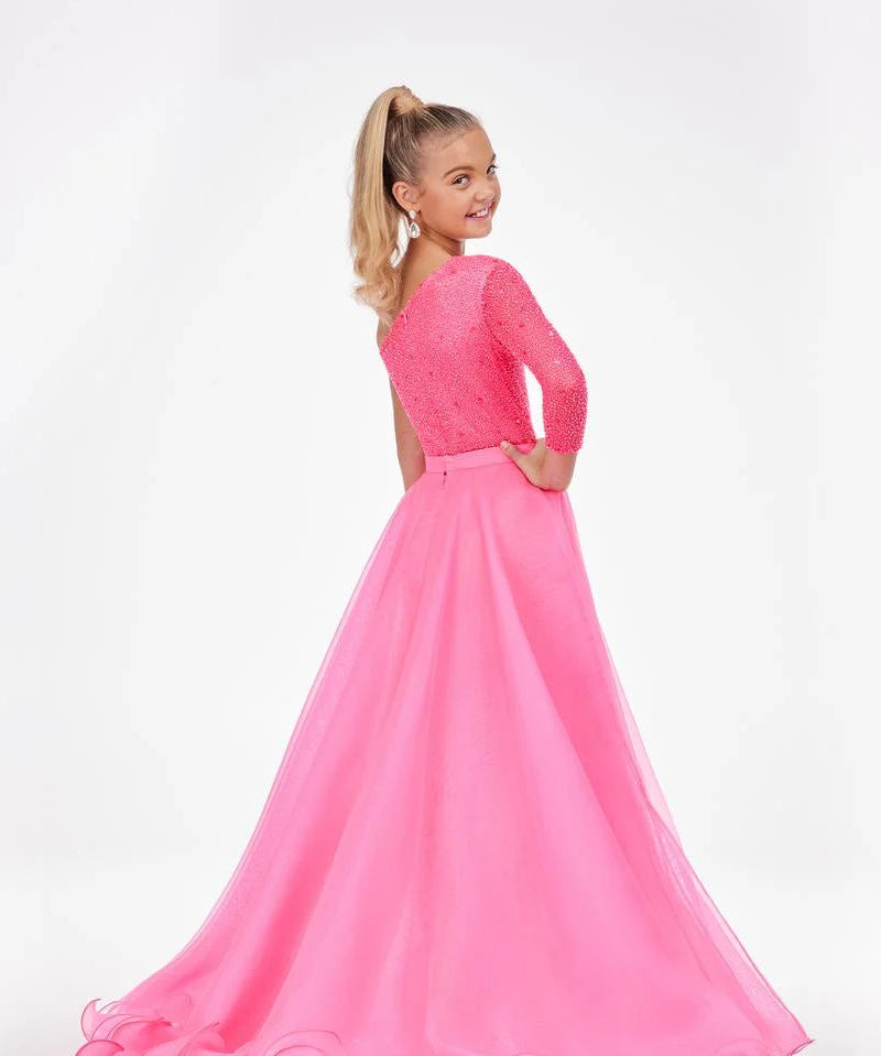 A young girl with blonde hair in a ponytail is shown from the back wearing a bright pink formal gown with a lace bodice and a flowing tulle skirt, set against a plain white studio background.