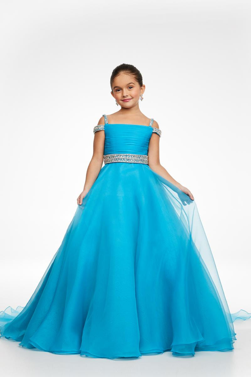 A young girl modeling a bright turquoise blue off-the-shoulder ball gown with a beaded silver waistband, photographed against a seamless white studio background.