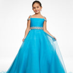 A young girl modeling a bright turquoise blue off-the-shoulder ball gown with a beaded silver waistband, photographed against a seamless white studio background.