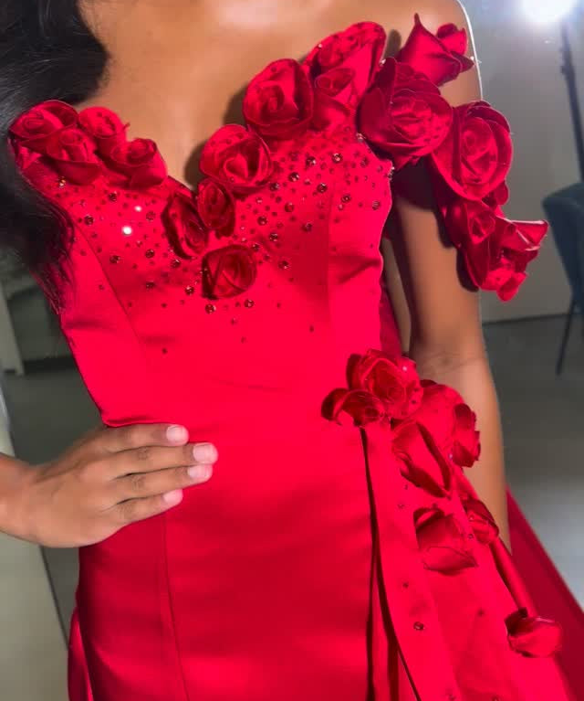 A close-up of a model wearing a vibrant red off-the-shoulder evening gown decorated with 3D rose appliqués and rhinestone embellishments.