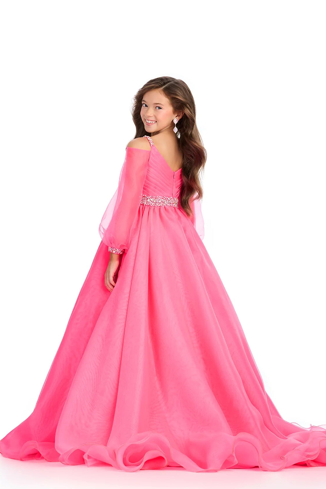 A young girl modeling a bright pink off-the-shoulder ball gown with a beaded waistband, photographed from the back against a solid white background.